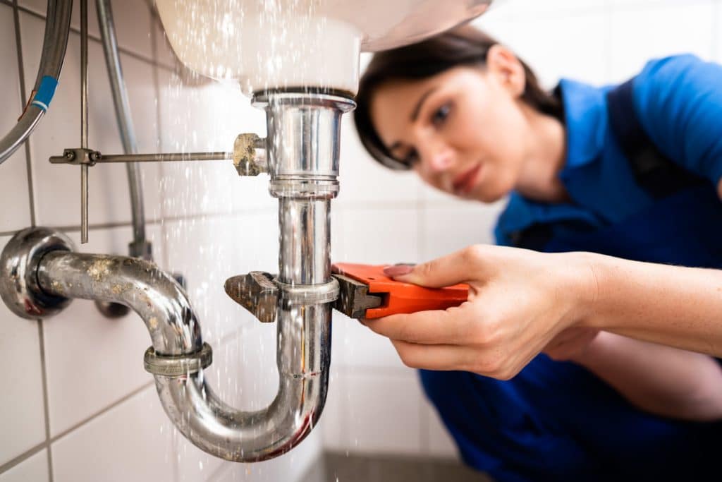 A plumber uses a pipe wrench to repair the chrome drain pipe beneath a sink while water sprays from the plumbing connection.