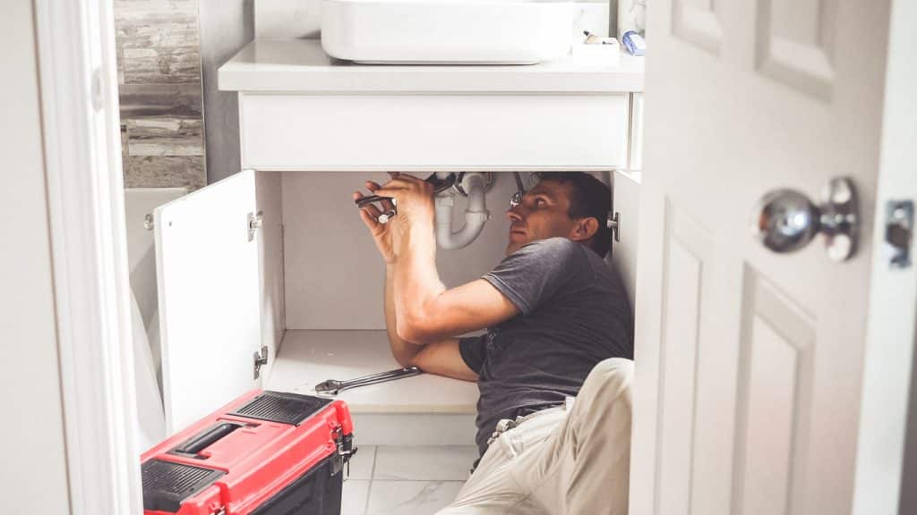 A plumber uses a wrench to repair pipes beneath a bathroom sink, with the cabinet doors open and a red toolbox nearby.